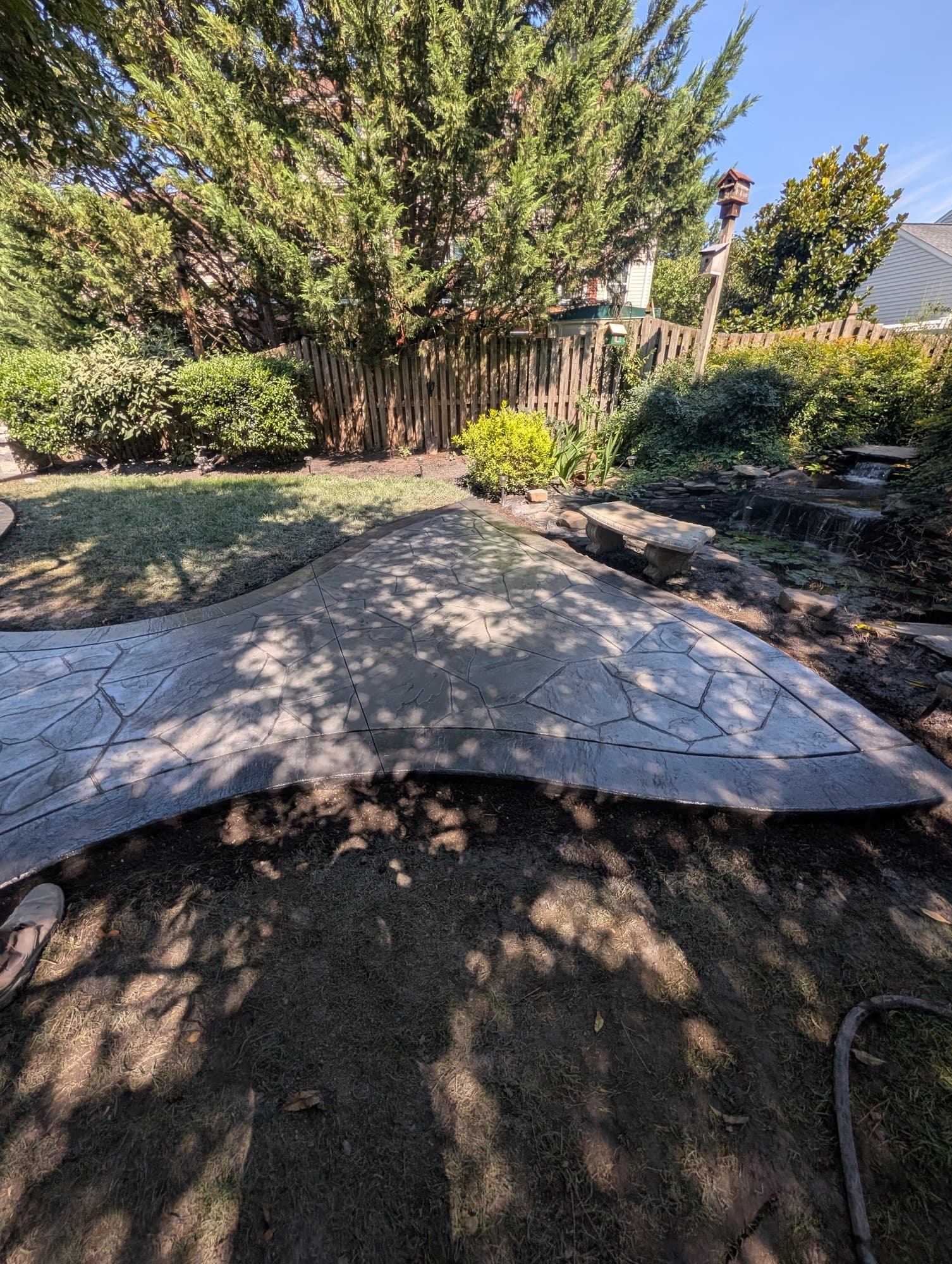 Winding stamped concrete walkway leading through a backyard toward a circular patio, with a garden pond and waterfall visible
