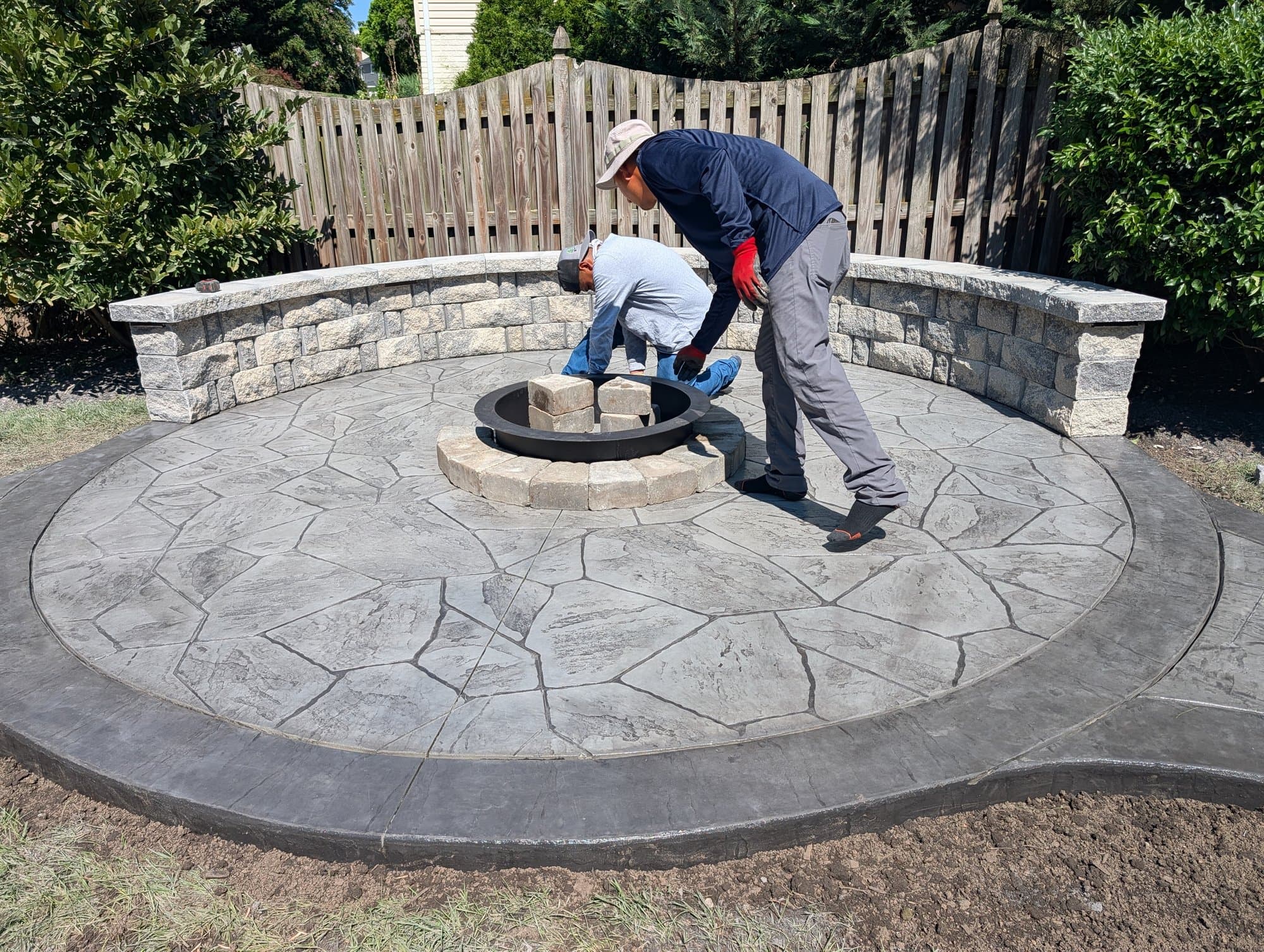 Workers installing a stone fire pit ring at the center of a circular stamped concrete patio