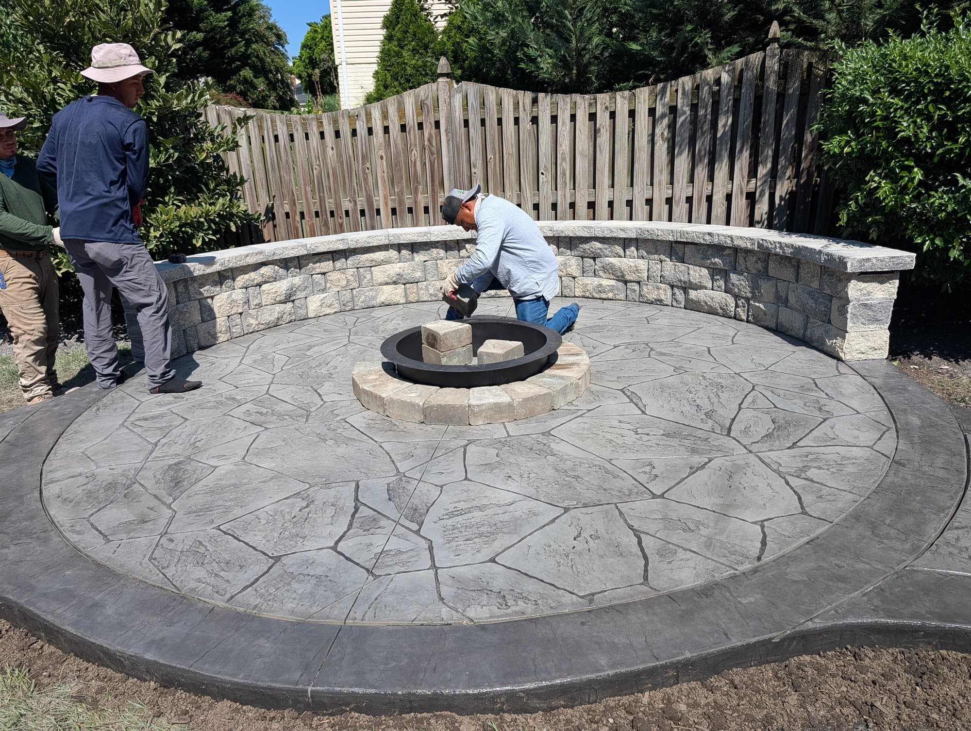 Workers installing a stone fire pit ring at the center of a circular stamped concrete patio, wider view